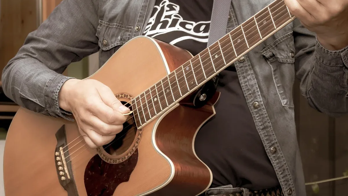 A person playing an acoustic guitar, wearing a gray denim jacket and a black t-shirt with white text.