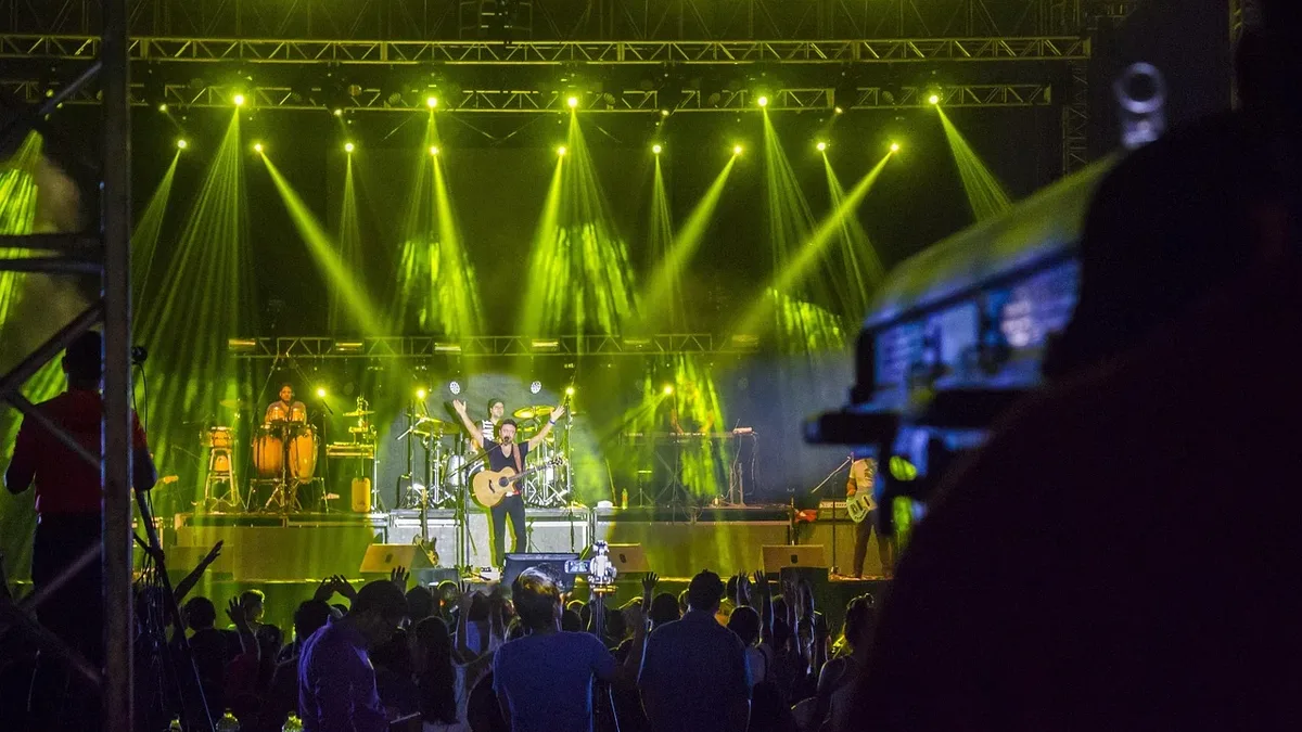 A live concert performance on a stage, featuring a band with a lead singer and guitarist, accompanied by a drummer and bassist, with a crowd of people in front of them.