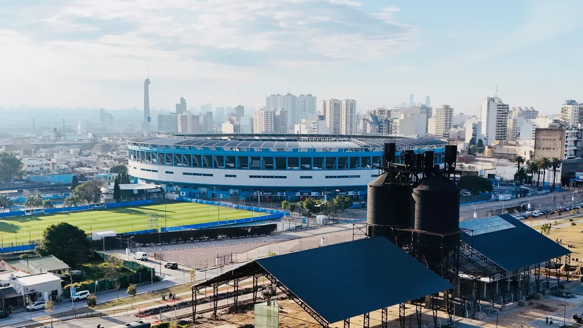 A large stadium with a blue and white exterior, a green field, and a city skyline in the background.